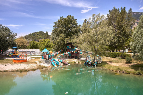Parco giochi con scivoli e stagno circondato da alberi al Camping Le Lac Bleu, Auvergne-Rhône-Alpes, Francia.