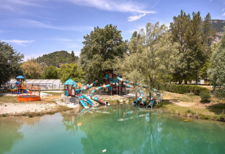 Speeltuin met glijbanen en een vijver, omgeven door bomen in Camping Le Lac Bleu, Auvergne-Rhône-Alpes, Frankrijk.