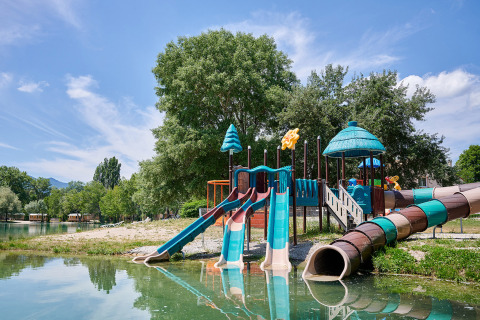 Playground with slides by a lakeshore, surrounded by trees, at Camping Le Lac Bleu in Auvergne-Rhône-Alpes, France.