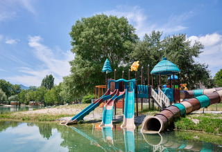 Playground with slides by a lakeshore, surrounded by trees, at Camping Le Lac Bleu in Auvergne-Rhône-Alpes, France.
