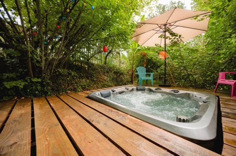 Jacuzzi exterior en terraza de madera con sillas y sombrilla, rodeado de árboles en Camping Le Pech Charmant, Francia.