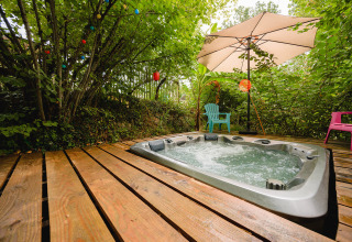 Bubbelbad buiten op houten terras, stoelen en parasol, omringd door groen in Camping Le Pech Charmant, Frankrijk.
