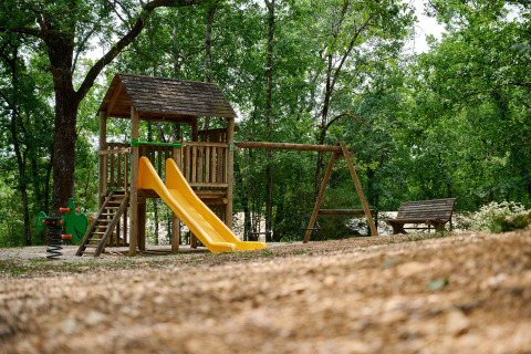 Parque infantil con tobogán, columpios y banco rodeado de árboles en Camping Le Pech Charmant, Francia.