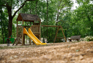 Outdoor playground with slide, swings, and bench surrounded by trees at Camping Le Pech Charmant, France.