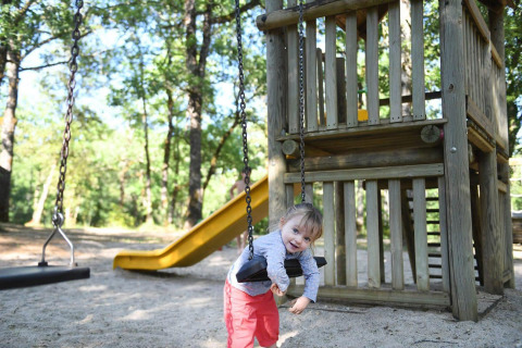 Niña pequeña con pantalones rojos juega en un columpio en parque infantil de madera y tobogán amarillo.