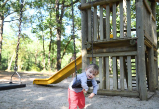 Small child in red pants playing on a swing at a playground with climbing tower and yellow slide in woods.
