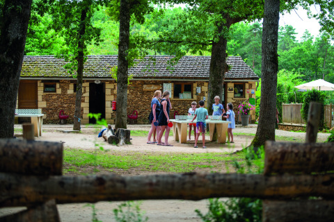 Familias juegan al ping-pong al aire libre en Camping Le Pech Charmant, rodeados de árboles y naturaleza.