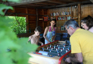 Gezin speelt tafelvoetbal in een houten chalet met boeken in Camping Le Pech Charmant, Nouvelle-Aquitaine.
