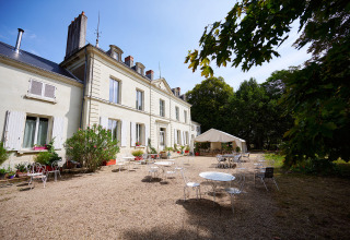 Udendørs siddeområde foran en elegant herregård på Camping Le Petit Trianon i Nouvelle-Aquitaine, Frankrig.