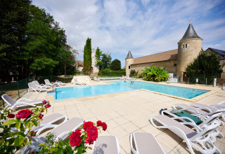 Udendørs swimmingpool med liggestole, blomster og historisk bygning på Camping Le Petit Trianon i Frankrig.