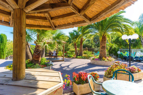 Zona exterior en Camping Leï Suves con palmeras, flores coloridas, asientos a la sombra y ambiente soleado.