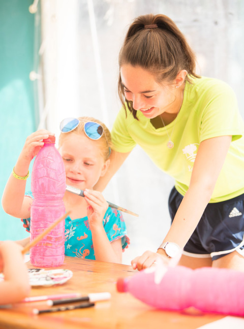 Una bambina dipinge una bottiglia di plastica rosa aiutata da un adulto al Camping Leï Suves in Provenza-Alpi-Costa Azzurra, Francia.