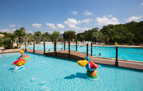 Outdoor pool at Camping Leï Suves, France with kids’ area, animal figures, a bridge, and lush palm trees.