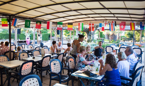 Restaurant im Freien im Camping Leï Suves, Frankreich, mit Gästen und internationalen Flaggen an der Decke.