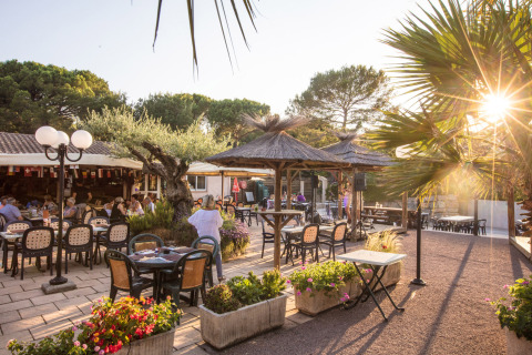 Zona de comedor al aire libre en Camping Leï Suves, Provenza, con palmeras, flores y comensales felices.