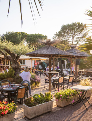 Zona de comedor al aire libre en Camping Leï Suves, Provenza, con palmeras, flores y comensales felices.