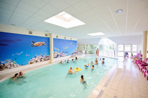 Piscine couverte avec familles et enfants, fresque murale d’océan et chaises longues roses alignées.