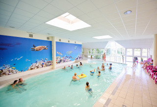 Piscine couverte avec familles et enfants, fresque murale d’océan et chaises longues roses alignées.