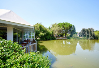 View of a pond and lush trees next to a terrace at Camping Les Bois du Bardelet, Centre-Val de Loire, France.