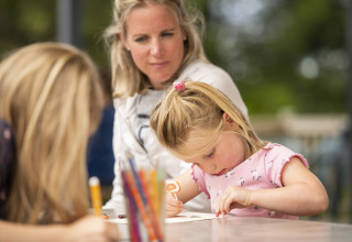 Una niña dibuja en una mesa bajo la mirada de una mujer en Camping Les Bois du Bardelet, Francia.