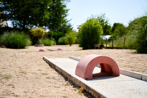 Campo de minigolf con obstáculo curvado, rodeado de arena y vegetación en Camping Les Bois du Bardelet, Francia.