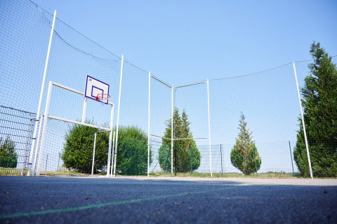 Outdoor basketball court with tall safety nets, green bushes, and clear blue sky at a holiday park.