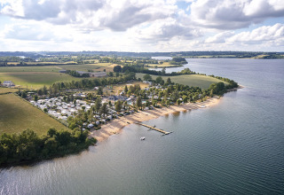 Aerial view of Camping Les Genêts holiday park by a lake in Occitanie, France, with a sandy beach and fields.