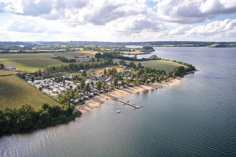 Aerial view of Camping Les Genêts holiday park by a lake in Occitanie, France, with a sandy beach and fields.