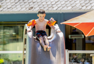 Boy wearing arm floaties sliding down a slide at Camping Les Genêts holiday park in Occitanie, France.