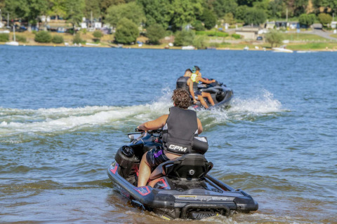 Personas montando motos acuáticas en el lago de Camping Les Genêts en Occitanie, Francia, rodeadas de naturaleza.