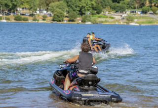 Folk kører på jetski på en sø ved Camping Les Genêts i Occitanie, Frankrig, omgivet af grønt landskab.