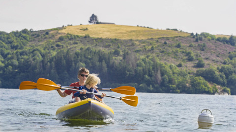 Two people paddling an inflatable kayak on a lake at Camping Les Genêts in Occitanie, France, with hills behind.