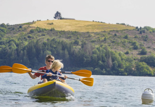 Two people paddling an inflatable kayak on a lake at Camping Les Genêts in Occitanie, France, with hills behind.