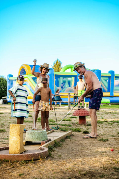 Familia jugando minigolf en Camping Les Genêts, parque vacacional en Occitanie, Francia, día soleado.