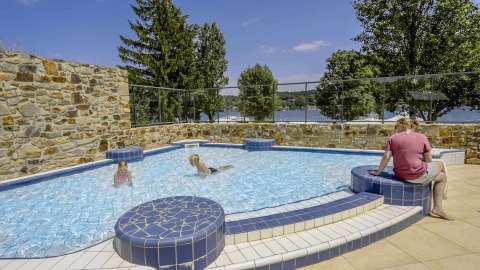 Piscina exterior en Camping Les Genêts, Francia, con niños jugando y vista al lago y naturaleza alrededor.