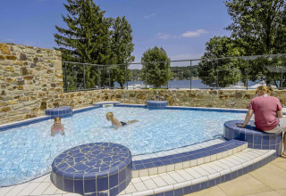 Outdoor swimming pool at Camping Les Genêts, France, with children playing and lake view in the background.