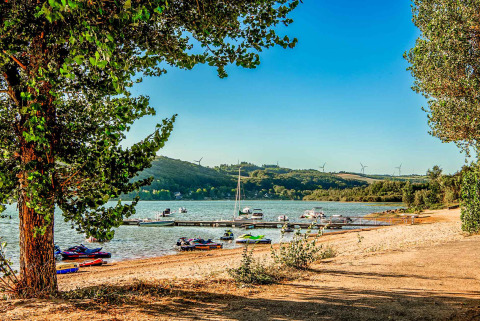 View of a lakeside at Camping Les Genêts in Occitanie, France, with boats, trees and wind turbines in background.