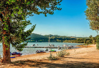 Vista sul lago al Camping Les Genêts in Occitania, Francia, con barche, alberi e turbine eoliche sullo sfondo.
