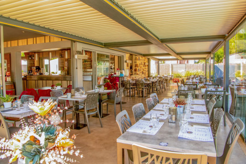 Restaurante moderno y luminoso con mesas preparadas y vistas al jardín en Camping Les Genêts, Occitanie, Francia.