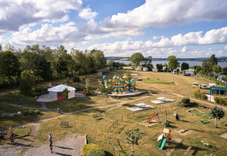 Vista aerea di Camping Les Genêts, parco vacanze in Occitania, Francia, con area giochi, verde e vista lago.