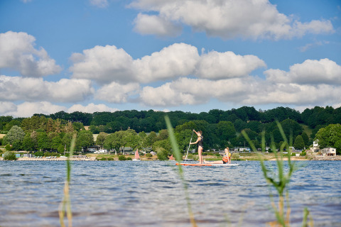 Family paddleboarding on a serene lake at Camping Les Genêts, surrounded by hills and cabins in Occitanie, France.