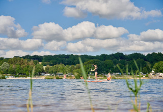 Family paddleboarding on a serene lake at Camping Les Genêts, surrounded by hills and cabins in Occitanie, France.