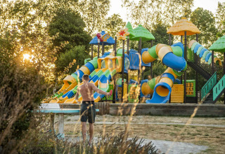 En mand står ved et bord foran en farverig legeplads med rutschebaner i Camping Les Genêts i Occitanie, Frankrig.