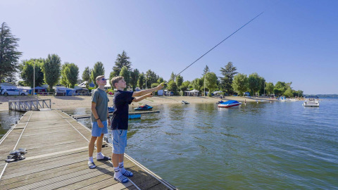 Two people fishing from a wooden pier at Camping Les Genêts holiday park in Occitanie, France, on a sunny day.