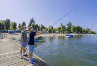 Two people fishing from a wooden pier at Camping Les Genêts holiday park in Occitanie, France, on a sunny day.