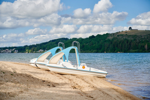 Dos botes de pedales en la orilla arenosa de un lago en Camping Les Genêts, Occitanie, Francia, con colinas verdes.