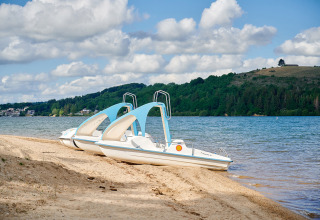 Two pedal boats on the sandy shore of a lake at Camping Les Genêts, Occitanie, France, with green hills.