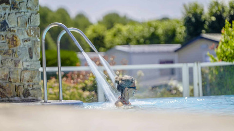 Mujer relajándose en la piscina bajo un chorro de agua en Camping Les Genêts, parque vacacional de Occitanie, Francia.