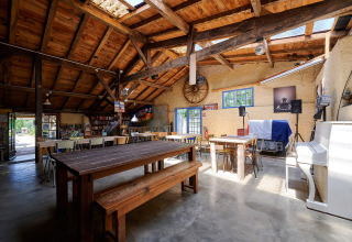 Salle commune lumineuse avec poutres apparentes, tables en bois, piano blanc au Camping Lestaubière, France.