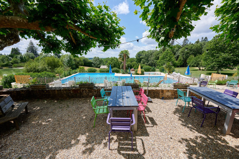 Area pranzo esterna con sedie colorate, tavoli e vista sulla piscina immersa nel verde a Camping Lestaubière, Francia.